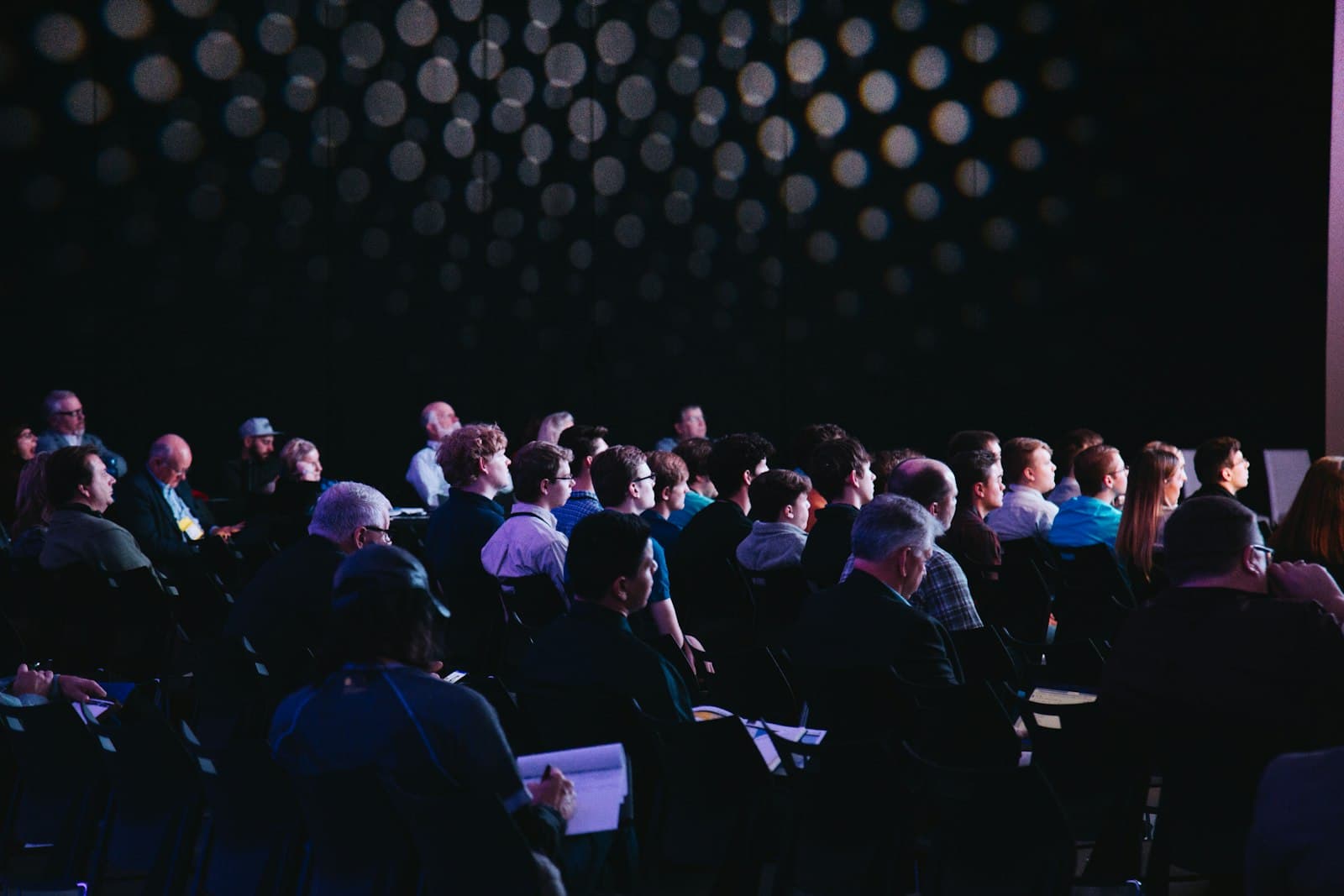 An audience seated in a dark hall, watching a speaker